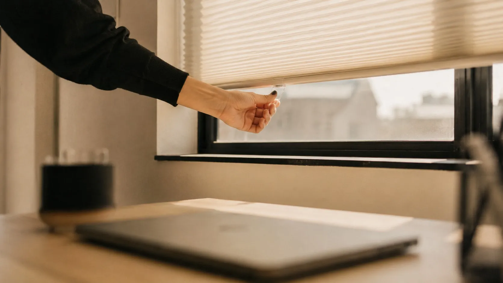 En hand drar ner en persienn framför ett kontorsfönster, en stängd laptop i förgrunden